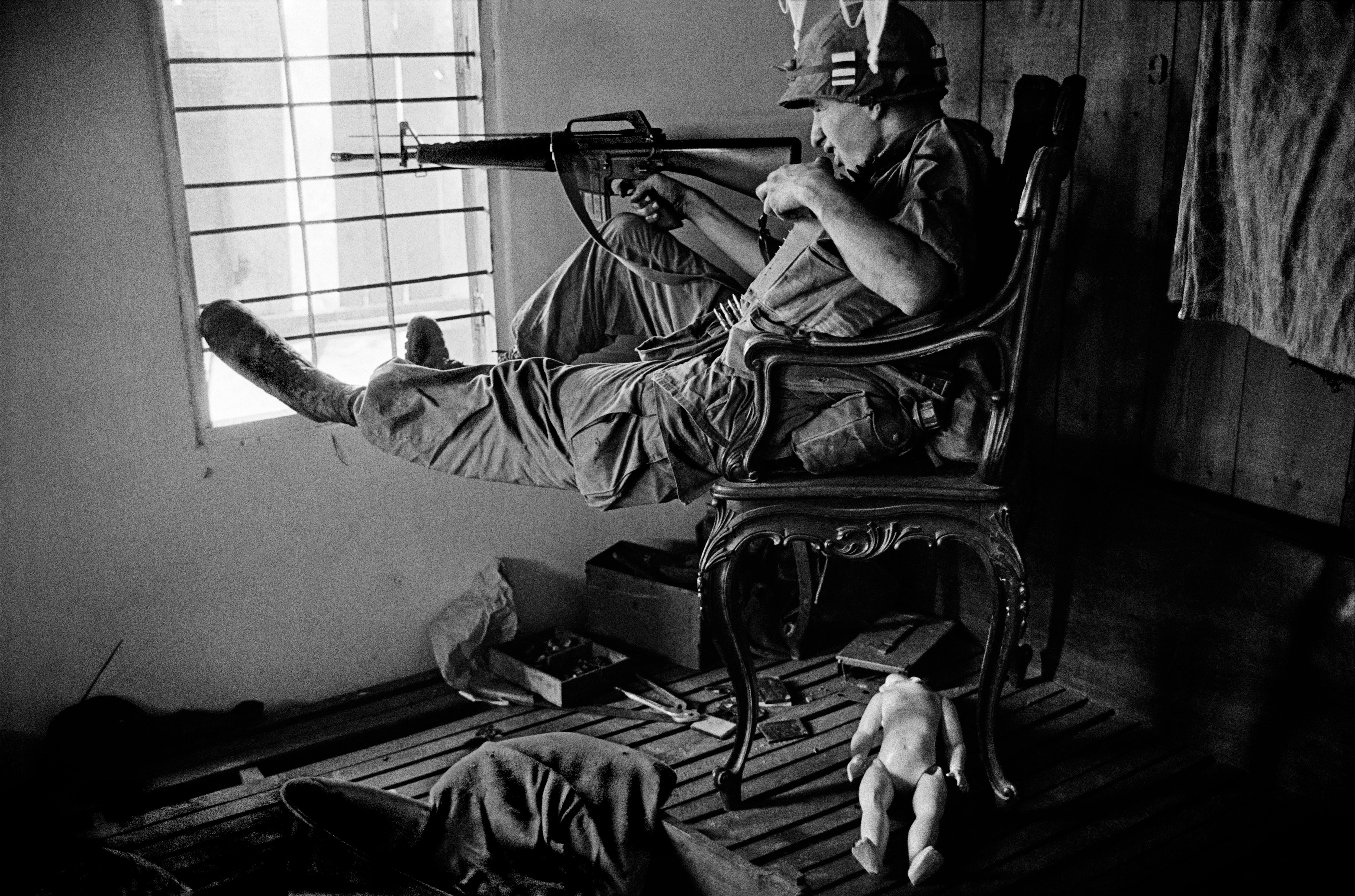 A soldier sitting, aiming a rifle out a window; a doll lies on the floor.
