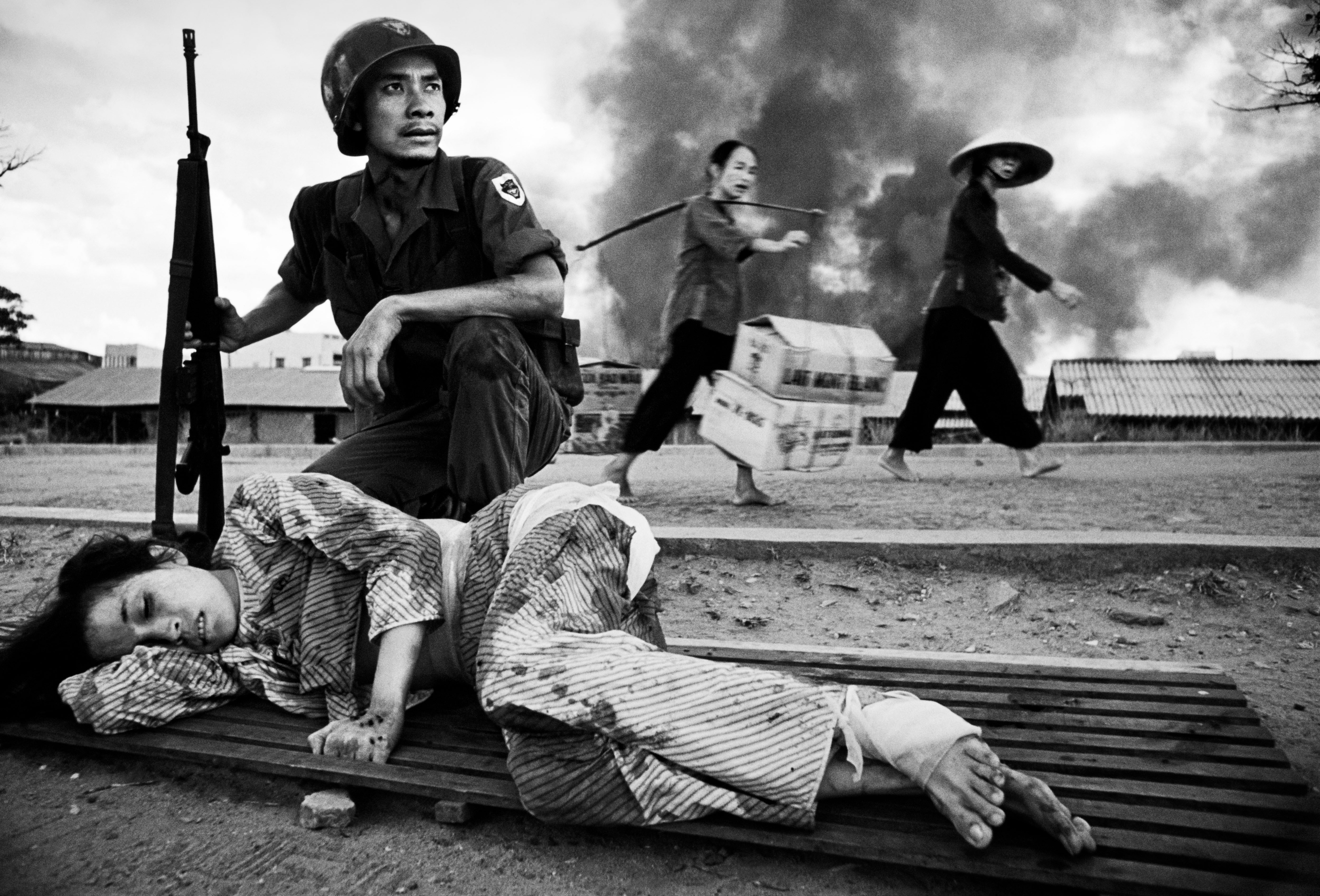 A wounded woman lying on a wooden plank during the Vietnam War, with a soldier and two women in the background.