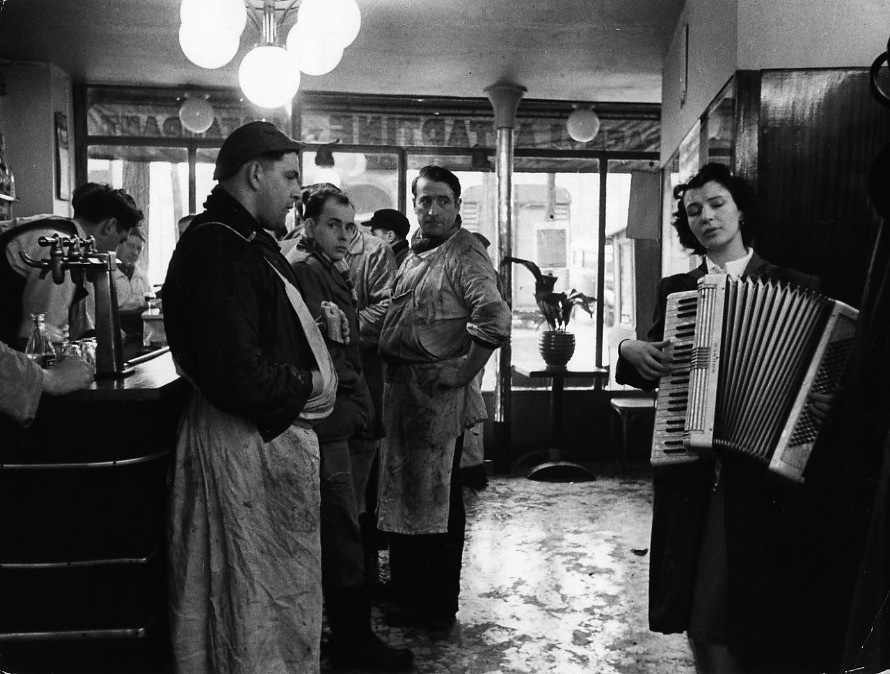 Black and white photo of a woman playing an accordion in a Parisian bar, butchers in the background.