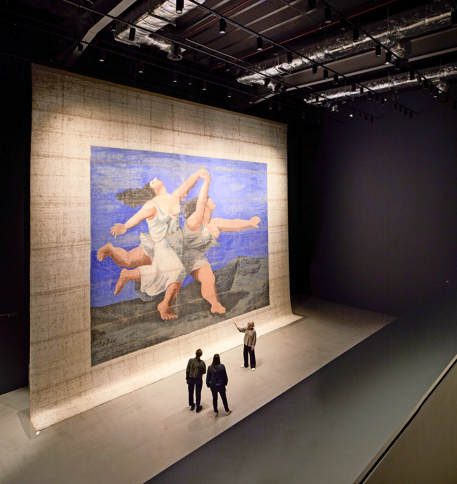 Large artwork depicting two women dancing, displayed in a gallery with viewers observing.