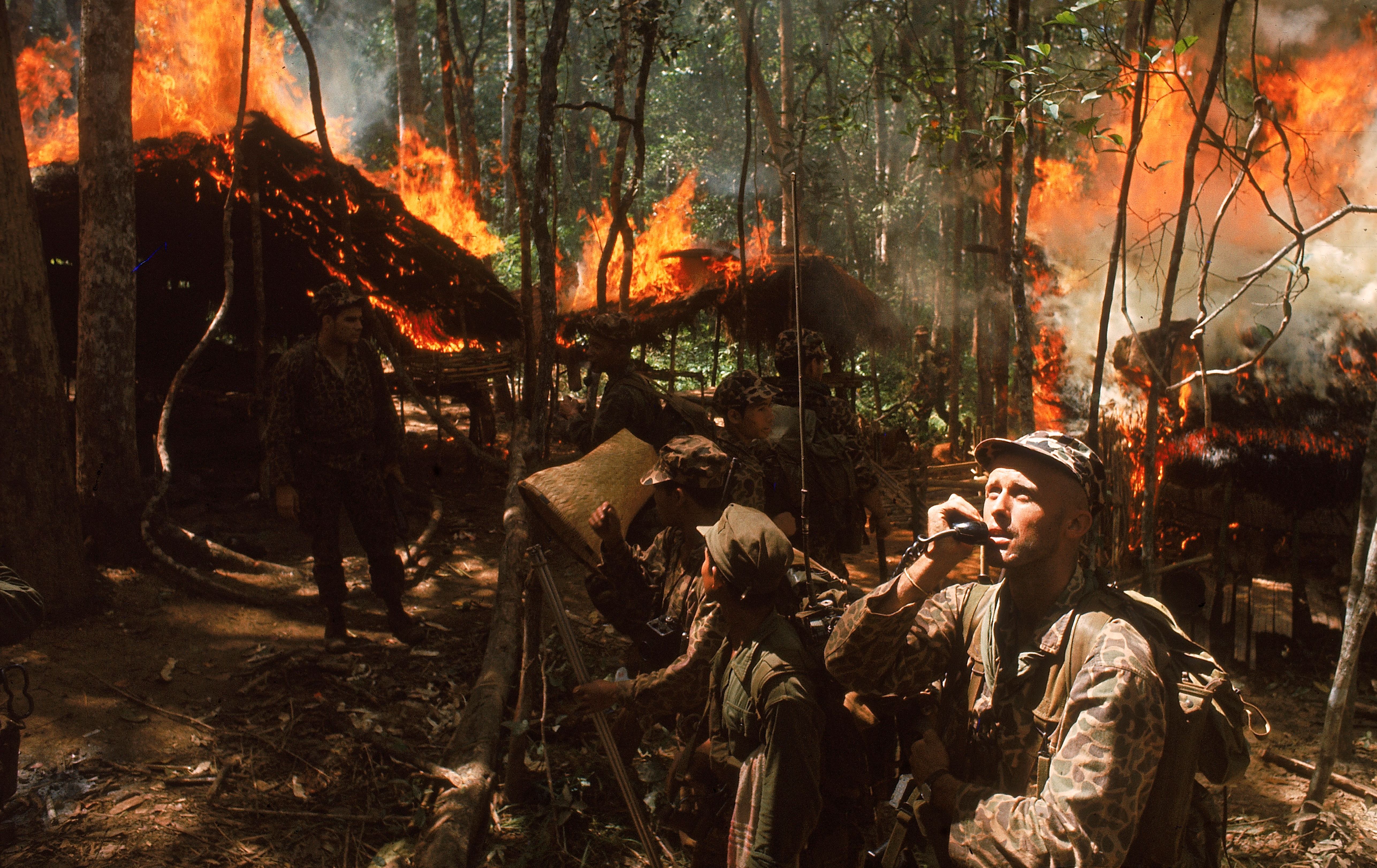 US Army Special Forces Capt. Vernon Gillespie using a radio while South Vietnamese soldiers burn a Viet Cong hideout.