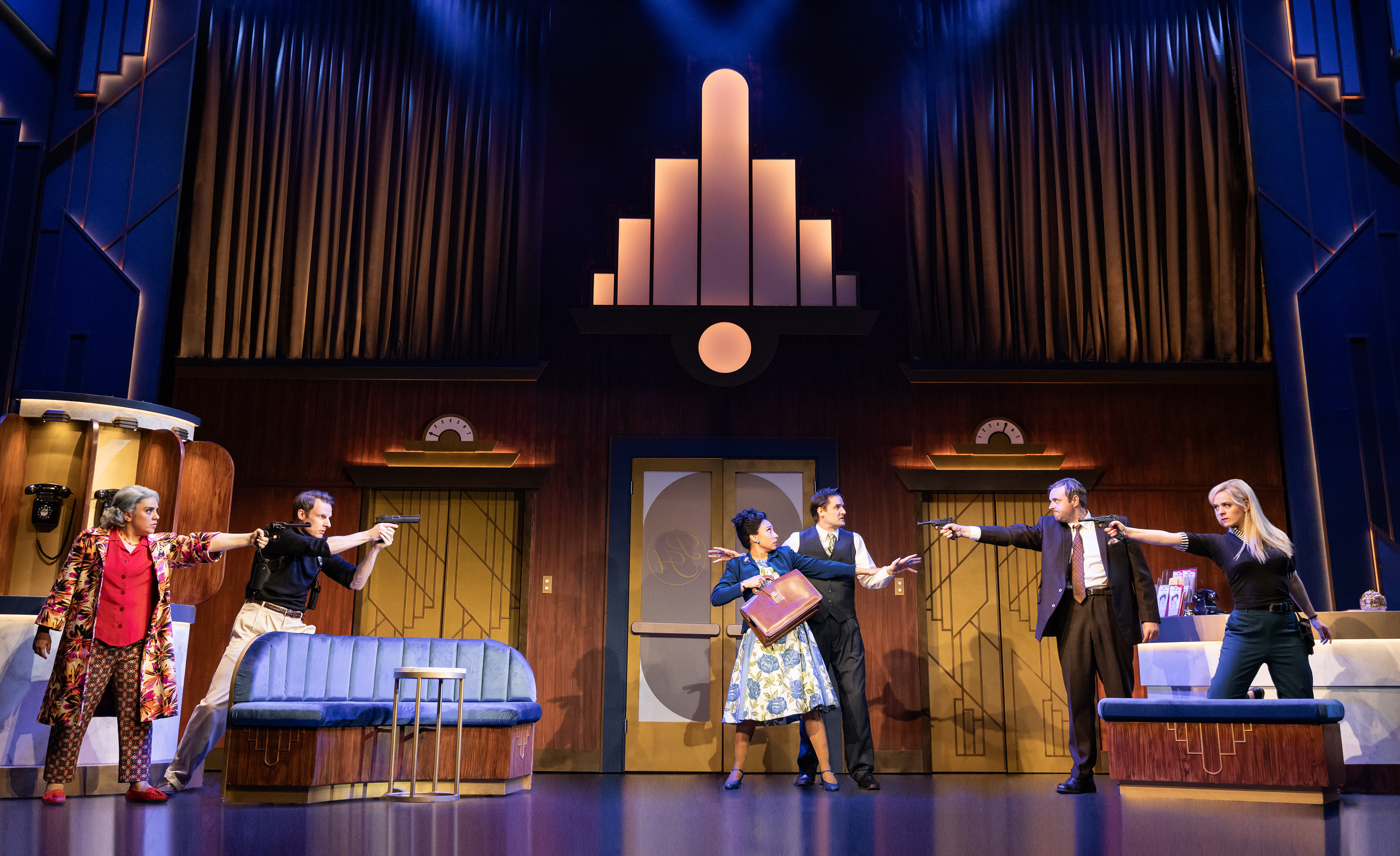 A scene from *The Comedy About Spies*, depicting six actors in a 1930s-style hotel environment, pointing guns at one another.